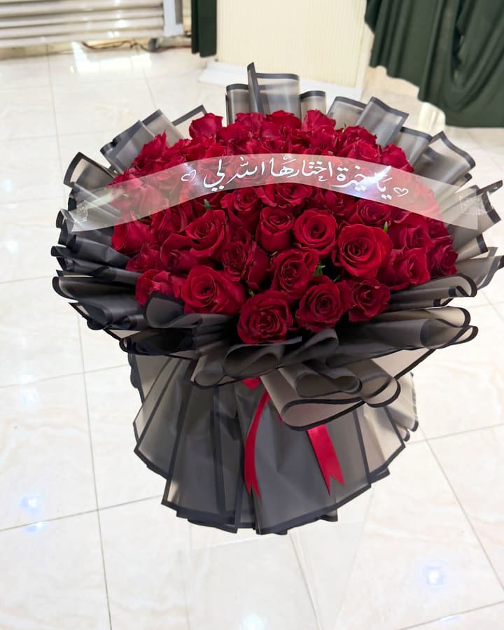 باقة كبيرة من الجوري الأحمر بتغليف أسود وشريط شفاف مكتوب عليه عبارة إهداء