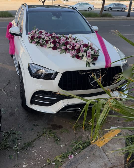سيارة مرسيدس بيضاء على كبوتها ورد وردي وعنابي وشريط تُل فوشي