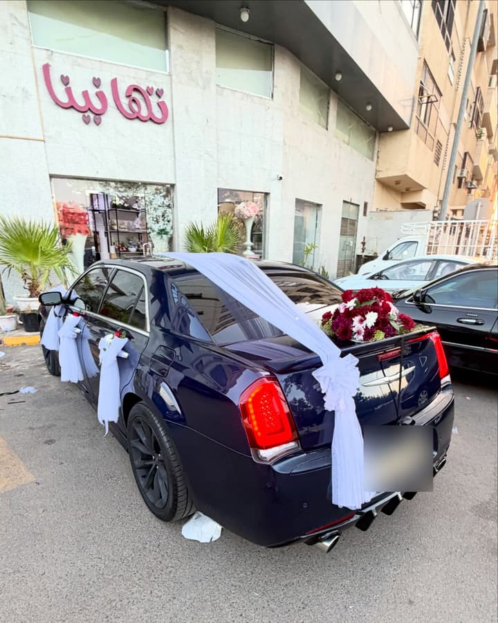 واجهة محل تهانينا باللوحة الوردية، وسيارة مزيّنة بتُل أبيض وورد أحمر أمامها