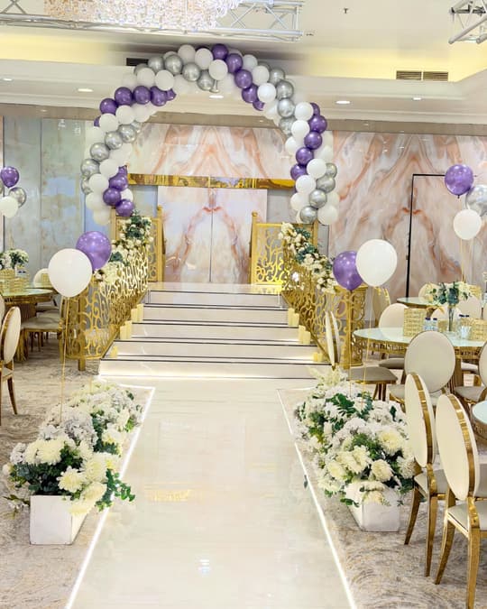ممر عروس أبيض تحفّه أحواض ورد أبيض وقوس بالونات بنفسجي وفضي